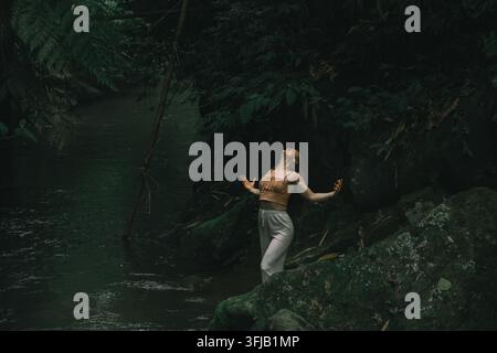 Femme debout sur un rocher moussu près d'une rivière de la jungle avec les bras ouverts et le dos cambré Banque D'Images