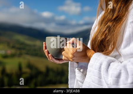 Femme tenant une tasse avec un fond de vallée montagneuse. Une femme en peignoir blanc tient une tasse grise devant une vallée montagneuse doucement floue sous un ciel bleu Banque D'Images