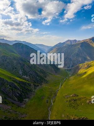 Une prise aérienne révèle une rivière sinueuse traversant une vallée montagneuse luxuriante au Kirghizistan. La lumière du soleil projette des ombres dramatiques sur le paysage verdoyant, Banque D'Images