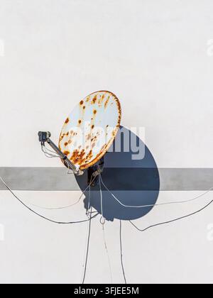 Ancien parabole satellite rouillée avec des fils projetant une ombre forte sur la façade d'un bâtiment Banque D'Images
