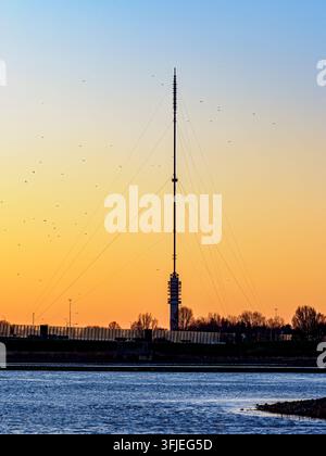 La tour Gerbrandy se dresse fièrement contre un ciel dégradé passant du bleu à l'orange vif, créant une silhouette frappante avec des oiseaux migrateurs volant au-dessus d'une rivière tranquille Banque D'Images