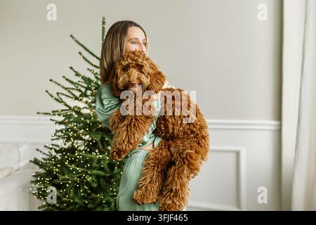 Close-up d'une jeune femme en pyjama en soie verte embrassant son adorable chien Labradoodle rouge. Belle scène de style de vie intérieur avec un sapin de Noël lumineux Banque D'Images