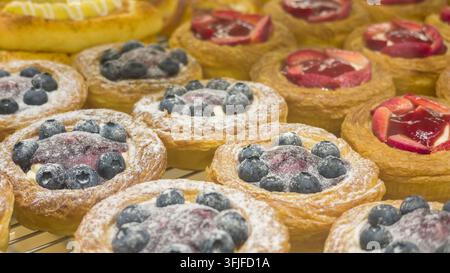 Close-up de pâtisseries danoises fraîchement cuites garnies de myrtilles, fraises, crème et sucre en poudre, exposées dans une boulangerie. Banque D'Images