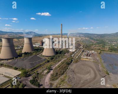 Centrale thermique industrielle remplissant une vallée rurale, avec trois tours de refroidissement, une cheminée centrale et des stocks de combustible. Une vue aérienne large met l'accent sur la chaleur Banque D'Images