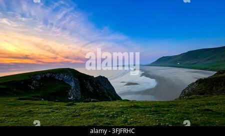 Le coucher de soleil pastel illumine la vaste plage de sable de Rhossili Bay, une falaise herbeuse encadrant la courbe de la côte. Péninsule de Gower, Pays de Galles. Banque D'Images