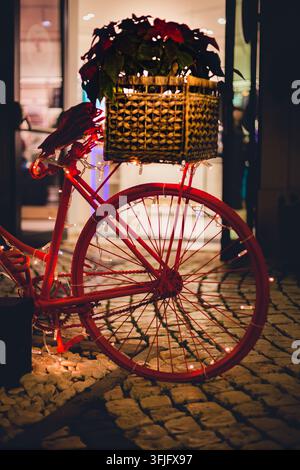 Gros plan d'un vélo vintage rouge décoré de guirlandes lumineuses et d'un panier de plantes de Poinsettia sur une rue pavée la nuit. Banque D'Images