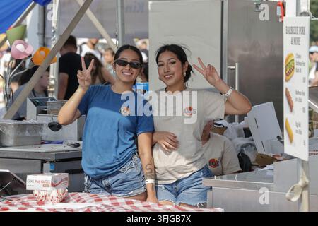 Bloomington, Minnesota, États-Unis - 13 juillet 2025 : Deux jeunes femmes asiatiques souriantes font des signes de paix au stand de nourriture de Lu lors d'un festival estival en plein air. Banque D'Images
