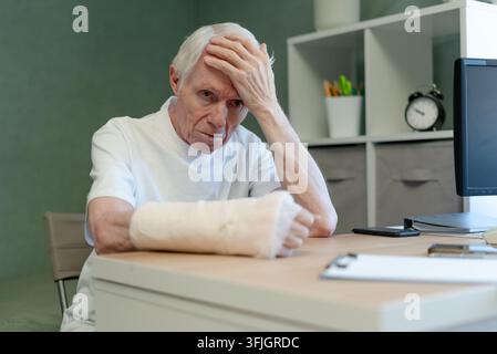 Homme âgé tenant sa tête au bureau, se sentant stressé et frustré. Homme senior lutte avec le travail de bureau pendant la convalescence d'une blessure au bras. Patient âgé fait face seul aux défis de santé dans l'espace de travail Banque D'Images