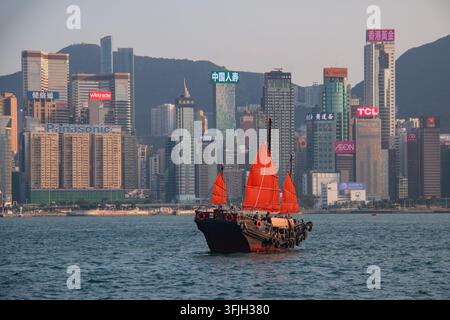 Bateau junk chinois traditionnel avec des voiles rouges vives traversant le Victoria Harbour devant l'horizon emblématique de l'île de Hong Kong au coucher du soleil Banque D'Images