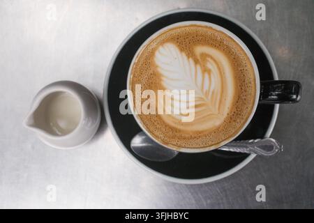 Un barista crée un art latte en forme de feuille complexe dans une tasse noire accompagnée d'une crème sur une table en métal dans un café. Banque D'Images