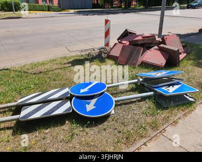Une collection de panneaux de signalisation routière et de matériaux de construction routière posés sur un accotement herbeux à côté d'un trottoir pavé et d'un carrefour. Dans le Banque D'Images
