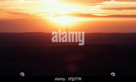 Coup d'œil aérien d'un coucher de soleil sur des collines ondulantes en Amérique avec une grande tour d'antenne de transmission s'élevant au-dessus du paysage forestier, ciel orange flamboyant, horizon paisible Banque D'Images