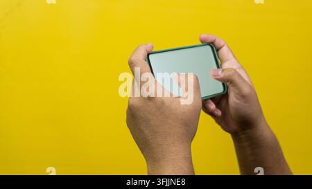 Deux mains d'hommes adultes saisissent un smartphone vierge horizontalement sur un fond jaune uni, démontrant des jeux mobiles actifs ou une saisie de texte rapide p Banque D'Images