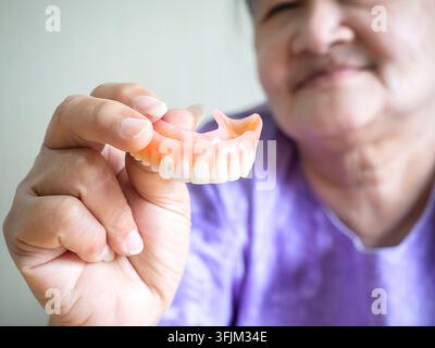 Ancienne femme asiatique souriant et tenant une dentier à la main. Les dentiers sont des dispositifs prothétiques conçus pour remplacer les dents manquantes et aider à mâcher Banque D'Images