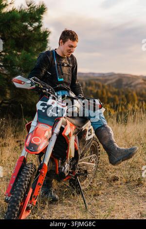 Jeune homme se repose sur sa moto tout-terrain au coucher du soleil Banque D'Images