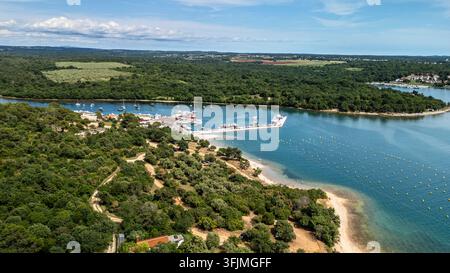 Panorama aérien d'une marina côtière avec des bateaux amarrés, une plage de sable et une eau turquoise encadrée par une forêt verte dense, Vabriga, comté d'Istrie Banque D'Images