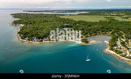 Vue aérienne d'une plage de sable côtière nichée à côté d'une forêt dense et d'un petit village, montrant des bateaux amarrés, une eau bleue claire, Vabriga, comté d'Istrie Banque D'Images