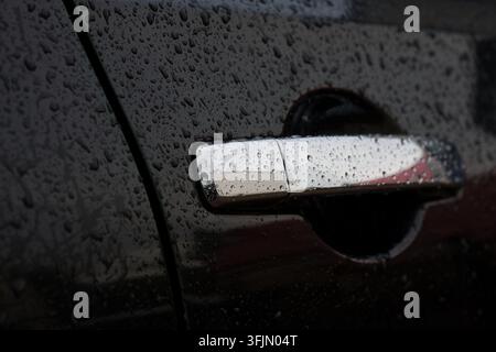 Grossissement d'une poignée de porte chromée brillante sur une carrosserie sombre recouverte de gouttelettes d'eau après la pluie. Banque D'Images