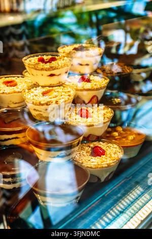 Desserts variés en couches dans des gobelets en plastique transparent exposés dans une vitrine réfrigérée de boulangerie. Plusieurs gobelets en plastique transparent contiennent diverses douceurs sucrées, notamment des parfaits avec des biscuits émiettés, de la crème et des fraises, des coupes de mousse au chocolat et des pecan au caramel Banque D'Images