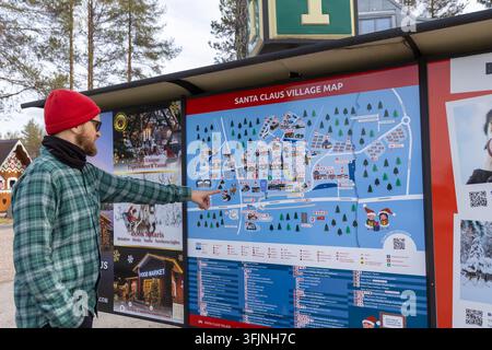 Éditorial : Rovaniemi, Finlande - 15 mai 2026 : Un homme barbu portant un bonnet rouge et une chemise à carreaux pointe une carte détaillée du Village du Père Noël Banque D'Images