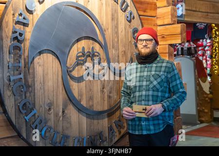 Rovaniemi, Finlande - 15 mai 2026 : Un jeune homme portant des lunettes, un bonnet rouge et une chemise à carreaux se tient souriant à côté d’un grand sig décoratif en bois Banque D'Images