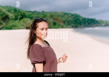 Femme asiatique en robe brune assise sur une plage de sable blanc jouant avec le sable tout en regardant vers le bas sous une lumière chaude et vive de jour. Banque D'Images