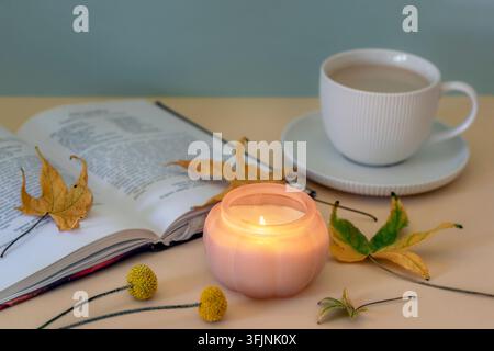 Bougie allumée, livre, tasse de café et feuilles d'automne tombées sur une table. Concept de maison confortable, gros plan. Banque D'Images