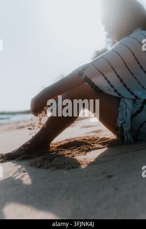 Vue de côté d'une femme assise sur une plage de sable regardant vers l'océan sous une lumière douce. Banque D'Images