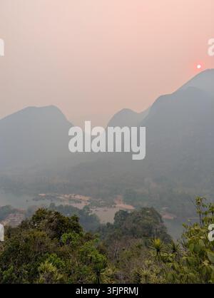 Vue panoramique au coucher du soleil mettant en scène un disque solaire rouge vif au-dessus de silhouettes de montagnes brumeuses et d'une rivière de vallée tropicale encadrée par des plantes vertes au premier plan. Banque D'Images