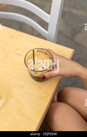 Close up d'une main tenant une boisson glacée au café freddo remplie de glaçons et de mousse de lait épaisse sur une table en bois dans un café grec en plein air Banque D'Images