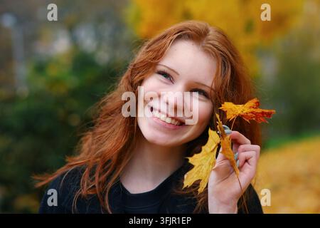 Jeune femme rousse souriante tenant des feuilles colorées dans un parc d'automne, exprimant la joie saisonnière, l'optimisme et la connexion avec la nature. Banque D'Images