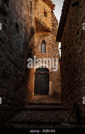Une ruelle historique étroite avec des murs en pierre rustique, une porte en bois et une arche à Tourrettes-sur-Loup. Une lumière chaude illumine l'ancienne architecture Banque D'Images