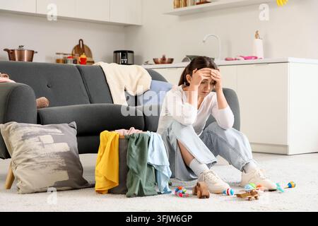 Épuisement parental et épuisement mental, mère fatiguée assise par terre avec du linge et des jouets dans un salon en désordre se sentant submergée par les tâches ménagères Banque D'Images