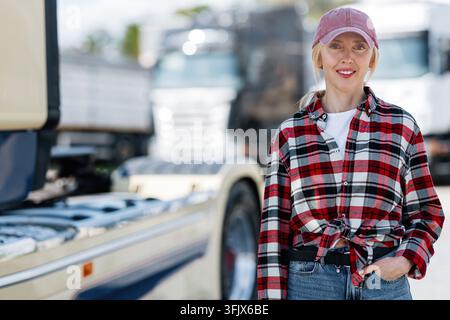 Une femme confiante portant une chemise à carreaux rouges et une casquette de baseball pose devant un grand semi-remorque par une journée ensoleillée Banque D'Images