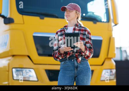Une femme portant une chemise à carreaux et une casquette de baseball tient une tablette numérique tout en travaillant à l'extérieur près d'un véhicule de transport lourd pendant la journée Banque D'Images