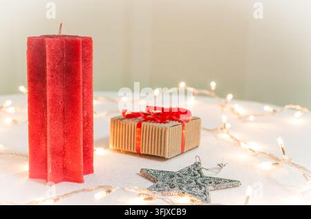 Une bougie en cire rouge en forme d'étoile se tient sur une surface légère à côté d'une boîte cadeau emballée, d'une étoile ornementale pailletée argentée et de guirlandes lumineuses chaudes. Banque D'Images