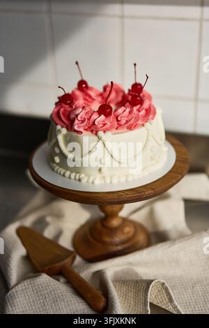 Petit gâteau de fête rond avec une garniture de crème au beurre blanche en forme de tuyau, un glaçage rose en rosettes et des cerises maraschino sur un support à gâteau en bois dans la lumière de la cuisine, avec Banque D'Images