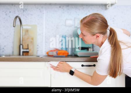 Femme souriante essuyant les armoires de la cuisine avec un chiffon, représentant les travaux ménagers quotidiens, la propreté et une maison saine. Banque D'Images