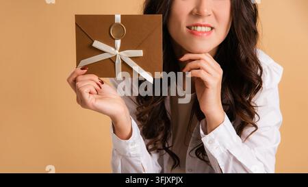 Femme souriante montrant une enveloppe cadeau artisanale avec sceau de cire et nœud ruban blanc sur fond beige Banque D'Images