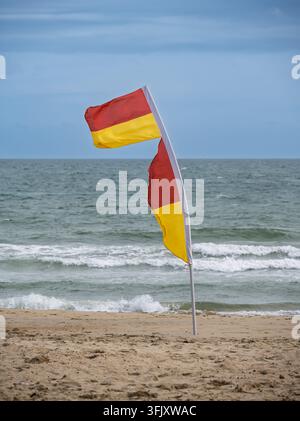 Un drapeau de sauvetage rouge et jaune flottant au vent sur une plage de sable, marquant la zone de baignade sûre contre les vagues de l'océan qui s'écrasent Banque D'Images