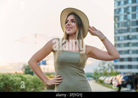 Jeune femme souriante touchant son chapeau de paille et regardant au loin en ville Banque D'Images