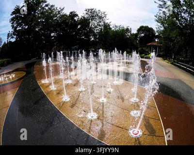 Plusieurs jets d'eau s'élèvent d'une place pavée décorative entourée d'arbres luxuriants et d'un kiosque dans un parc public. Banque D'Images