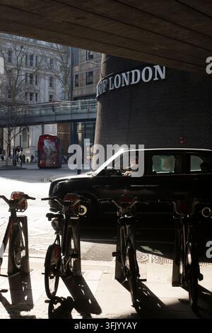 Un chauffeur manœuvre un taxi noir sous le passage souterrain du Musée de Londres sur London Wall, traversant la ville devant des vélos garés. Banque D'Images