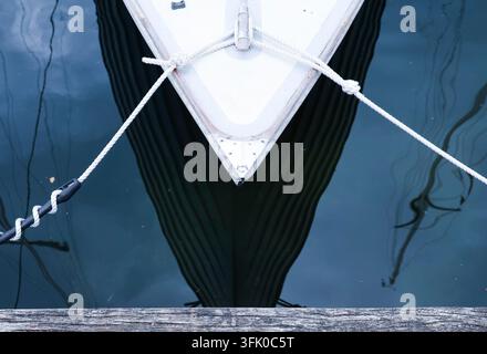 Arc de bateau blanc avec des cordes d'amarrage reflétant dans l'eau encore bleue d'un port Banque D'Images
