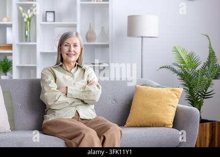 Femme mûre aux cheveux gris et aux bras croisés assise sur un canapé confortable dans un salon moderne, souriant à l'objectif, incarnant les concepts de bonheur, de détente et de vie indépendante à la maison Banque D'Images
