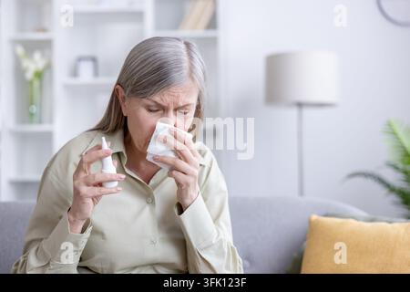 Femme âgée aux cheveux gris utilisant un spray nasal et tenant un mouchoir contre son nez sur un canapé, gérant les symptômes du rhume ou d'allergie et cherchant un soulagement à l'intérieur Banque D'Images