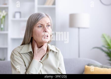 Femme âgée à la maison avec une gorge douloureuse, touchant son cou et grimçant de douleur, montrant des symptômes de maladie, de douleur, de difficulté à avaler et une inquiétude pour sa santé Banque D'Images
