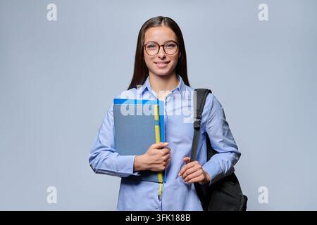 Portrait d'une jeune étudiante heureuse portant un sac à dos et tenant des classeurs et des cahiers bleus. Femme souriante aux lunettes regardant la caméra dans un bureau Banque D'Images
