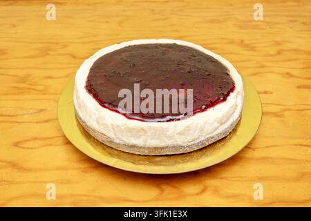 Cheesecake rond entier recouvert d'une compote de fruits rouges brillante, posé sur une planche à gâteau dorée sur une surface en bois chaud. Banque D'Images
