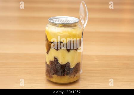 Dessert de gâteau mangue en couches avec une génoise au chocolat dans un récipient en boîte transparente et une cuillère sur une surface en bois. Banque D'Images